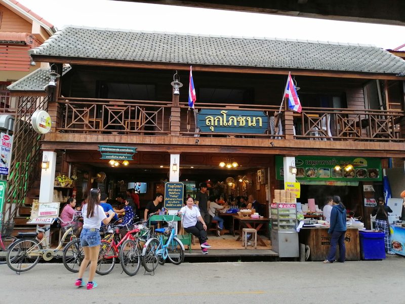 หน้าร้านลุกโภชนา เรือนไม้สองชั้นมีป้ายชื่อร้านสีเขียวและธงชาติที่ระเบียง